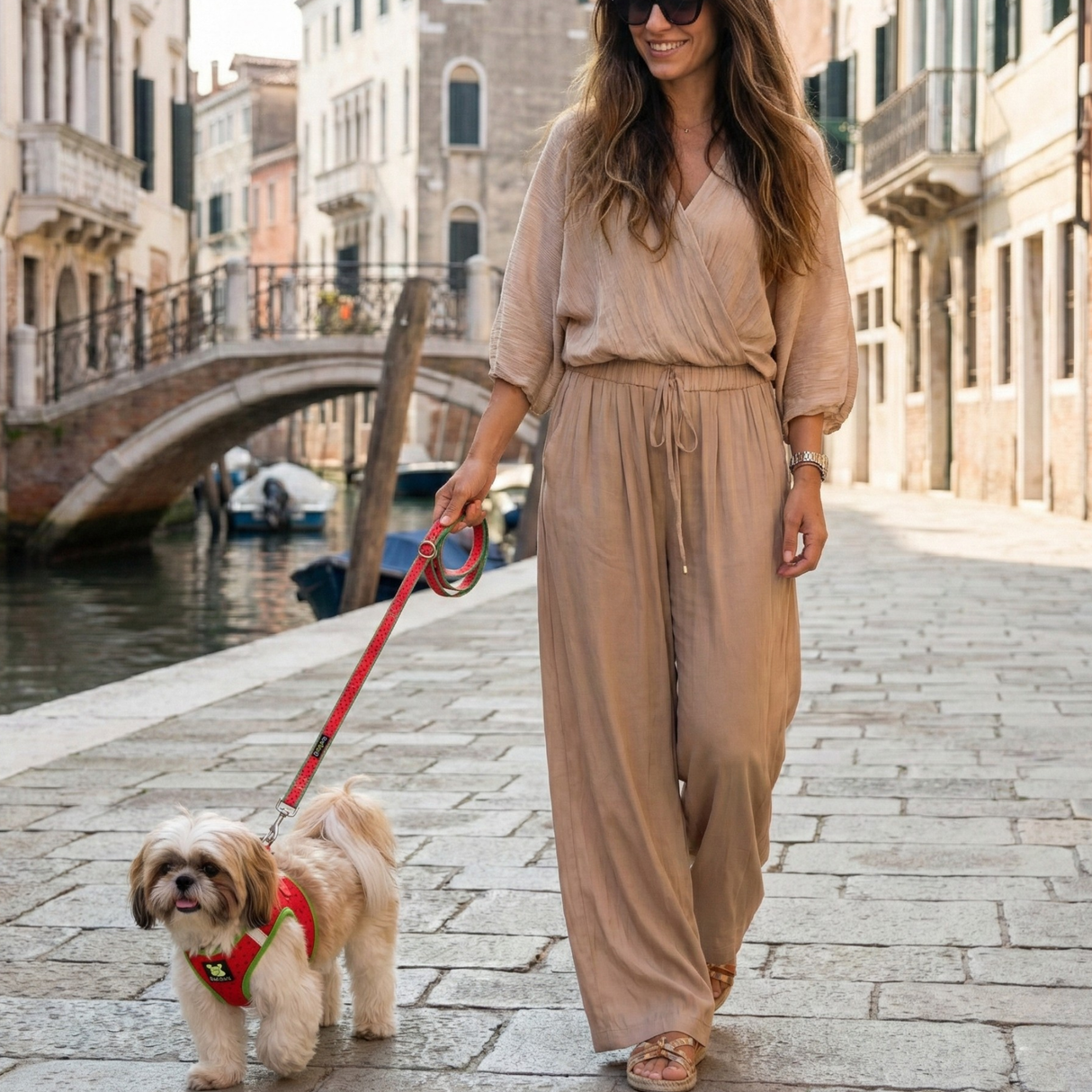 A woman in a beige jumpsuit walking a small Shih Tzu by a canal in an Italian town. The small dog is wearing an EcoBark mesh step-in dog harness and matching leash in the watermelon pattern.