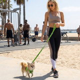 A woman in athletic wear walking an xxs fluffy chihuahua wearing a lime green EcoBark mesh step-in dog harness and matching leash on a sandy path at Muscle Beach