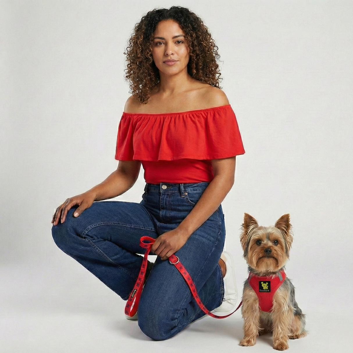 A woman wearing a red top and jeans kneeling next to a tiny Yorkshire Terrier wearing a red EcoBark mesh step-in dog harness and matching leash.