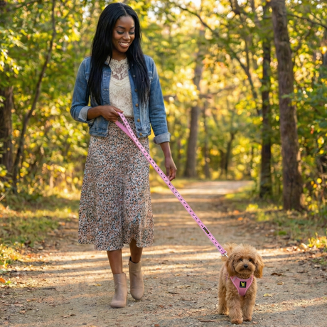 A smiling woman in a denim jacket walking her brown Toy Poodle on a wooded trail using an xs Baby Pink EcoBark harness and matching patterned leash